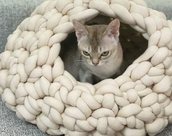 以下が含まれることがあります： 太い編み込み状の布チューブで作られた、ライトベージュ色のチャンキーニット製猫用ベッド。洞窟のような円形の構造で、中央の大きな開口部の中に、尖った耳を持つ小さなライトブラウンの猫が収まっています。