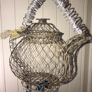 May include: A silver wire teapot with a decorative handle made of a silver spring. The teapot is decorated with blue and green glass beads hanging from the bottom.