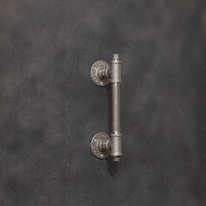 Könnte beinhalten: Ein silberner Türgriff im Industriestil aus Rohrverbindungen. Der Griff hat zwei runde Flansche mit Zahlen darauf.