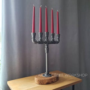Op de afbeelding: Een industriële kandelaar met vijf rode kaarsen. De kandelaar is gemaakt van grijze metalen buizen en fittingen, gemonteerd op een houten voet. De tekst "ART CRAFT WORKSHOP" is zichtbaar op de voet. Unieke woondecoratie.