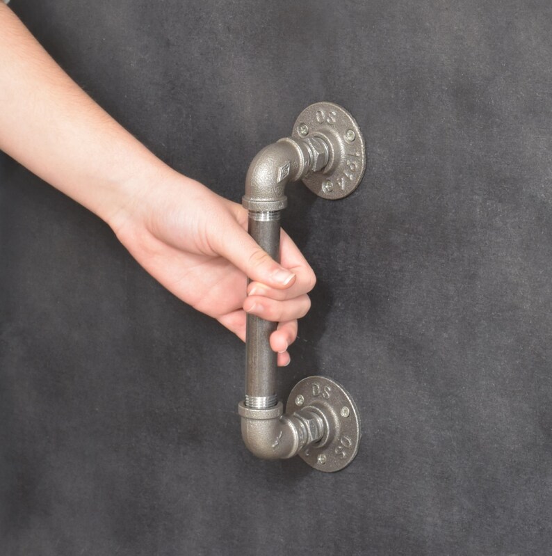 May include: A black metal pipe door handle with a round flange on each end. The handle is attached to a wall.