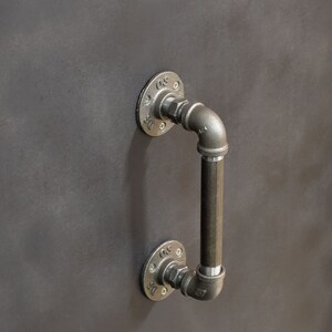 May include: A black metal industrial style door handle with a pipe design. The handle is attached to a wall with two flanges.