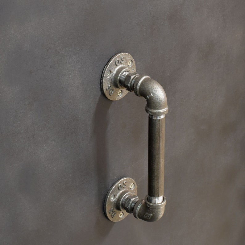 May include: A black metal industrial style door handle with a pipe design. The handle is attached to a wall with two flanges.