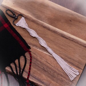 以下が含まれることがあります： ねじれたロープのデザインとタッセルが付いた白いマクラメキーホルダー。キーホルダーはシルバーのキーリングに取り付けられています。