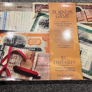 May include: A vintage board game box with a brown background and a title that reads "Business Game: A Mining Transport Strategy Game". The box features a collage of old stock certificates with images of trains and ships. The text "Jeu d'Affaires: Un Jeu de Stratégie du Transport de Minerai" is also visible on the box.
