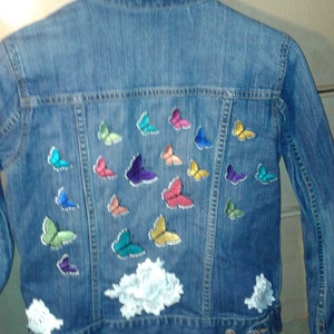 Puede incluir: Una chaqueta vaquera azul con parches de mariposas de colores y aplicaciones de encaje blanco.
