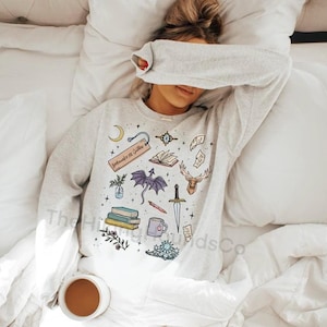 May include: Light gray sweatshirt with a whimsical design featuring a crescent moon, open books, a purple dragon, a deer head, a sword, and other fantasy-themed illustrations. The text "Bookmarks & Quill" is visible on a bookmark. A coffee cup is in the foreground.