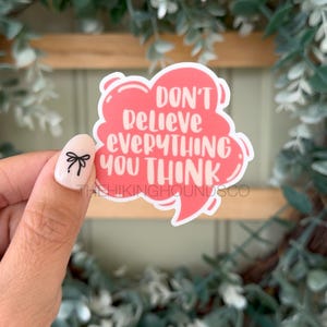 May include: A pink sticker in the shape of a speech bubble with the words "DON'T BELIEVE EVERYTHING YOU THINK" in white. The sticker is held by a hand with a bow design on the fingernail.