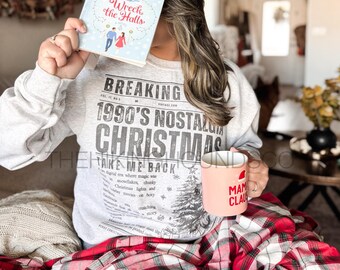May include: A person holding a book titled "Wreck the Halls" by Tessa Bailey, with a light blue cover. They are wearing a gray sweatshirt with the text "BREAKING 1990'S NOSTALGIA CHRISTMAS TAKE ME BACK". A pink mug with "MAMA CLAUS" is also visible.