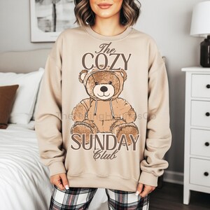 May include: Beige sweatshirt featuring a brown teddy bear wearing a hoodie. The text "The COZY SUNDAY Club" is printed in brown. The sweatshirt has long sleeves and a crew neck. The design is centered on the front of the garment.