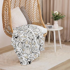 May include: A white throw blanket with a repeating pattern of ghosts and stars. The ghosts are drawn in a simple, cartoon style and are surrounded by small stars. The blanket is draped over a wicker chair.