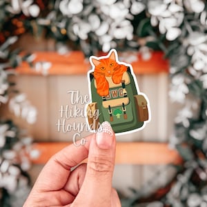 May include: A sticker featuring an orange cat resting on a green backpack. The backpack has brown accents and the letters "WG". The sticker also has the text "The Hiking Hounds Co."