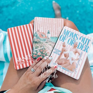 May include: Three rectangular e-reader cases with light pink and white vertical stripes. One case features a coastal lighthouse scene, another displays the text "SANDY SUN KISSED," and the third shows two figures with the text "MOMS OUT OF ORDER."
