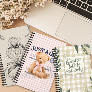 May include: Three spiral-bound notebooks with floral and teddy bear designs. One notebook features a large iris illustration, another has a pink gingham pattern with a teddy bear and text, and the third has a green gingham pattern with the text "Reminder: talk to God daily."