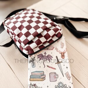 May include: A burgundy and white checkered crossbody bag with a black strap. A white phone case with fantasy-themed illustrations, including a dragon, sword, and books, is partially visible. The text "THE HINGING ROUNDS CO" is printed on the case.