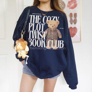 May include: Navy blue sweatshirt with the text "THE COZY PLOT TWIST BOOK CLUB" and a teddy bear graphic wearing a plaid shirt. A small teddy bear keychain hangs from a black purse strap. The sweatshirt is paired with a blue and white checkered skirt.