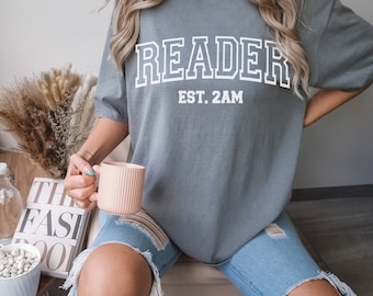 May include: A gray t-shirt with the text "READER EST. 2AM" in white. The person wearing the shirt is holding a pink mug.