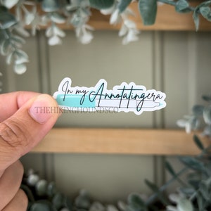 May include: A sticker with a teal highlighter and the text "In my annotating era" in black cursive font. The sticker is on a wooden surface with green plants in the background.