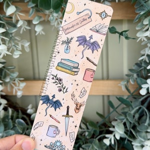 May include: A bookmark with a light brown background and a variety of illustrations, including a dragon, a deer, a book, a sword, a teacup, and a crown. The text "Bookmarks are not Quitters" is printed on the bookmark.