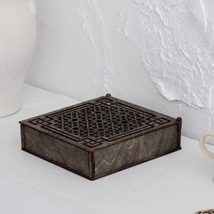 Op de afbeelding: Een decoratieve houten doos met een geometrisch roosterontwerp, een kleine beige theekop en een bijpassende onderzetter. Een grote witte vaas met twee handvatten is ook aanwezig. De items zijn gerangschikt op een witte plank tegen een getextureerde witte muur.