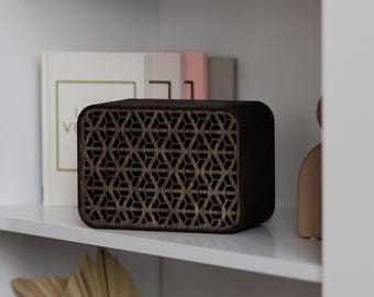 May include: A dark brown wooden speaker with a geometric pattern on the front. The speaker is sitting on a white shelf with other decorative items.