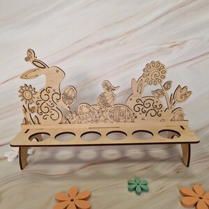 May include: Wooden Easter egg holder with a bunny, bird, and floral design. The holder has eight slots for eggs.