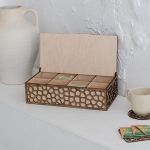 Op de afbeelding: Een houten theedoos met een decoratief geometrisch patroon op de voorkant, met theezakjes. De doos is open en toont de compartimenten. Een witte keramische mok en een kleine onderzetter zijn ook aanwezig.