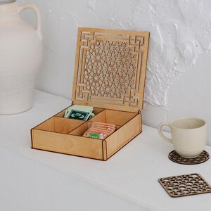 Könnte beinhalten: Eine Holzteebox mit dekorativem, durchbrochenem Deckel, unterteilt in vier Fächer. Die Box enthält Teebeutel. Eine weiße Tasse steht auf einem passenden Untersetzer. Eine dekorative Vase befindet sich im Hintergrund.