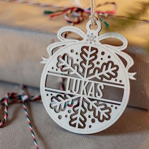 Puede incluir: Adorno navideño de madera con forma de copo de nieve con el nombre "LUKAS" recortado en el centro. El adorno cuelga frente a un regalo envuelto. Está atado con hilo natural.