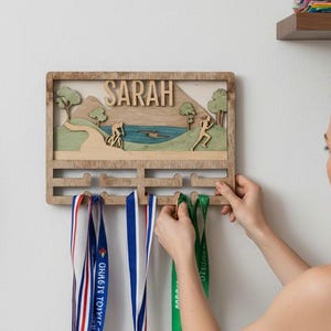 May include: Wooden medal display with the name "SARAH" and a landscape scene. The display has several hooks holding medals with colourful ribbons. A woman is seen hanging a medal on the display. A bicycle wheel is visible on the left.