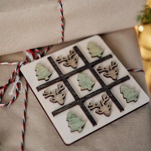 Könnte beinhalten: Ein hölzernes Tic-Tac-Toe-Spiel mit Weihnachtsmotiv. Das Spielbrett zeigt ein Raster mit Spielfiguren in Form von Rentieren und Weihnachtsbäumen. Das Brett ist aus hellem Holz mit dunkelbraunen Gitterlinien. Das Spiel liegt auf verpackten Geschenken.