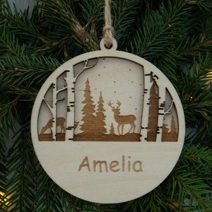 Pu&ograve; includere: Un ornamento natalizio rotondo in legno con un design a laser di una scena di foresta invernale. L'ornamento presenta due alberi, un cervo e il nome "Amelia" inciso sul fondo.