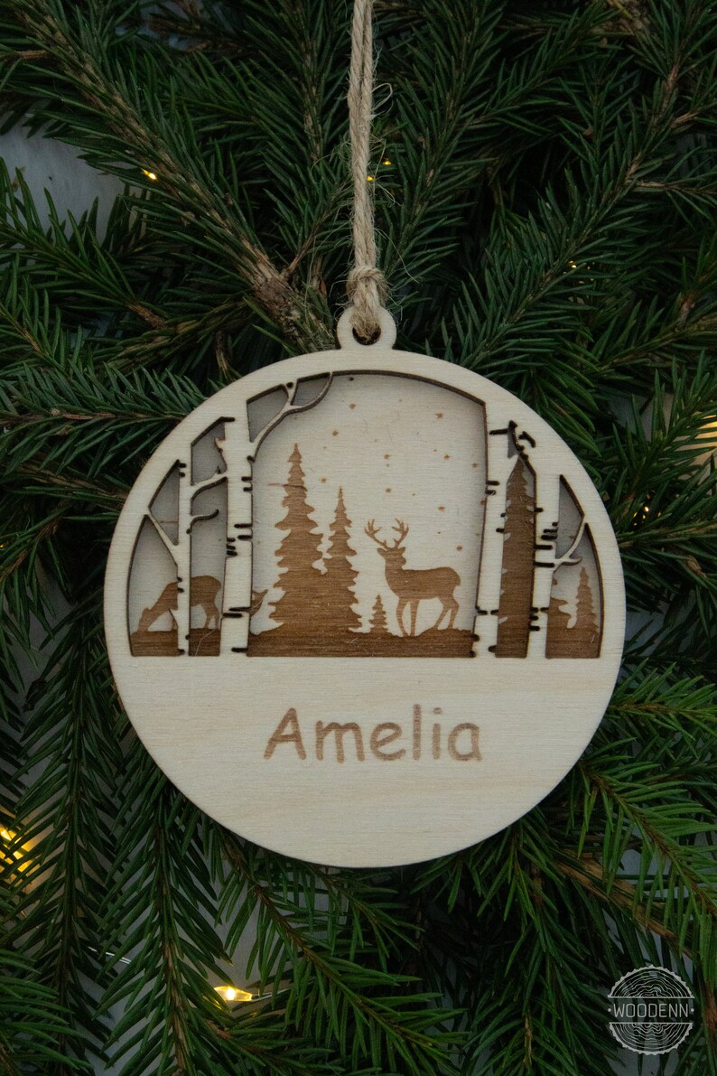 Pu&ograve; includere: Un ornamento natalizio rotondo in legno con un design a laser di una scena di foresta invernale. L'ornamento presenta due alberi, un cervo e il nome "Amelia" inciso sul fondo.