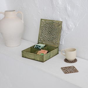 May include: A green wooden tea box with a decorative geometric design on the lid, open to reveal tea packets. A white ceramic mug sits on a matching coaster. A white vase is in the background.