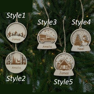 Pu&ograve; includere: Cinque stili diversi di addobbi natalizi in legno con nomi personalizzati. Ogni addobbo presenta un design a sfera di neve con diverse scene invernali, tra cui una casa, un camion, un pupazzo di neve e un cervo. Gli addobbi sono etichettati con i nomi "James", "Amelia" e "Noel".