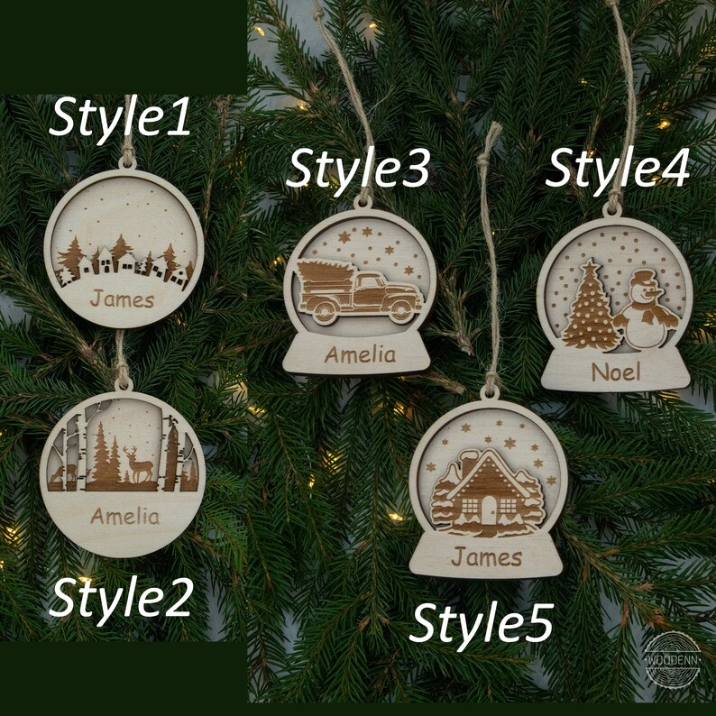 Pu&ograve; includere: Cinque stili diversi di addobbi natalizi in legno con nomi personalizzati. Ogni addobbo presenta un design a sfera di neve con diverse scene invernali, tra cui una casa, un camion, un pupazzo di neve e un cervo. Gli addobbi sono etichettati con i nomi "James", "Amelia" e "Noel".