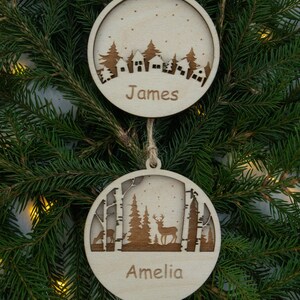 Pu&ograve; includere: Due decorazioni natalizie in legno con una scena invernale. L'ornamento superiore presenta un villaggio innevato con il nome "James" inciso sotto. L'ornamento inferiore rappresenta una scena di bosco con un cervo e il nome "Amelia" inciso sotto.