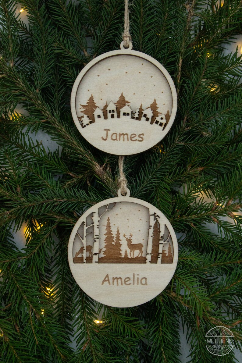 Pu&ograve; includere: Due decorazioni natalizie in legno con una scena invernale. L'ornamento superiore presenta un villaggio innevato con il nome "James" inciso sotto. L'ornamento inferiore rappresenta una scena di bosco con un cervo e il nome "Amelia" inciso sotto.