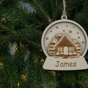 Pu&ograve; includere: Un ornamento natalizio in legno a forma di sfera di neve con una casa all'interno. L'ornamento &egrave; personalizzato con il nome "James".