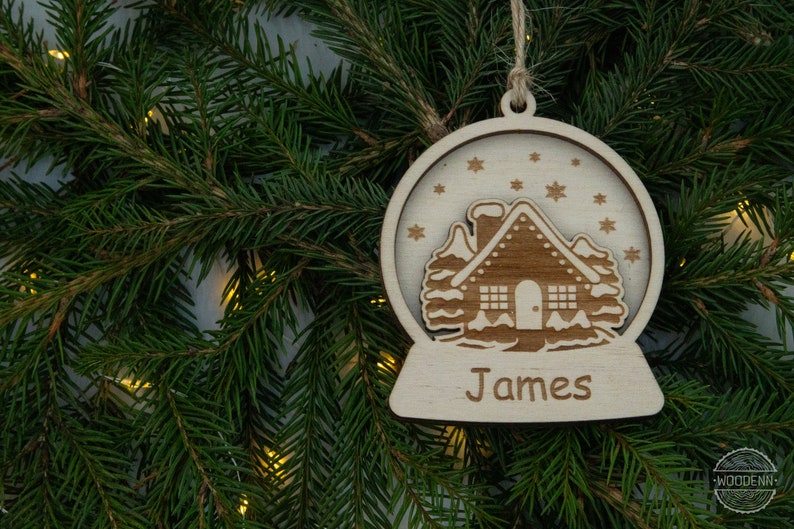 Pu&ograve; includere: Un ornamento natalizio in legno a forma di sfera di neve con una casa all'interno. L'ornamento &egrave; personalizzato con il nome "James".