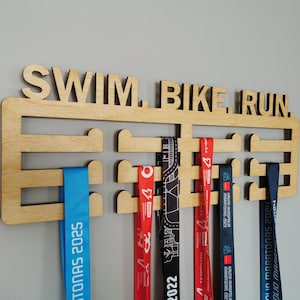 May include: Wooden medal display with the words "SWIM. BIKE. RUN." cut out at the top. Several colorful ribbons hang from the display, including blue, red, and black ribbons with text and dates.