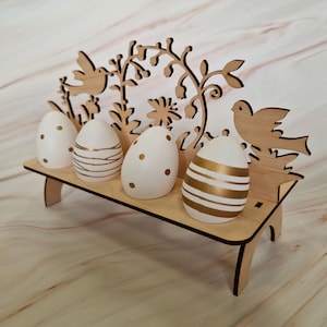 Op de afbeelding: Houten paaseierenhouder met een bloemenmotief en twee vogels. De houder heeft vier vakjes voor eieren, en de eieren zijn versierd met gouden stippen en strepen.