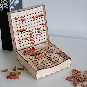 Könnte beinhalten: Holz-Schiffe-Versenken-Spiel mit einem Klappdeckel. Das Spielbrett ist hellbraun mit einem Raster aus Löchern und Buchstaben und Zahlen an den Seiten. Rote und braune Holzschiffteile sind auf dem Brett platziert.