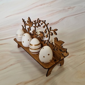 Può includere: Porta uova di legno per Pasqua con un design di uccello e fiori. Il porta uova ha cinque slot per le uova ed è dipinto di bianco. Le uova sono decorate con strisce dorate e pois bianchi.