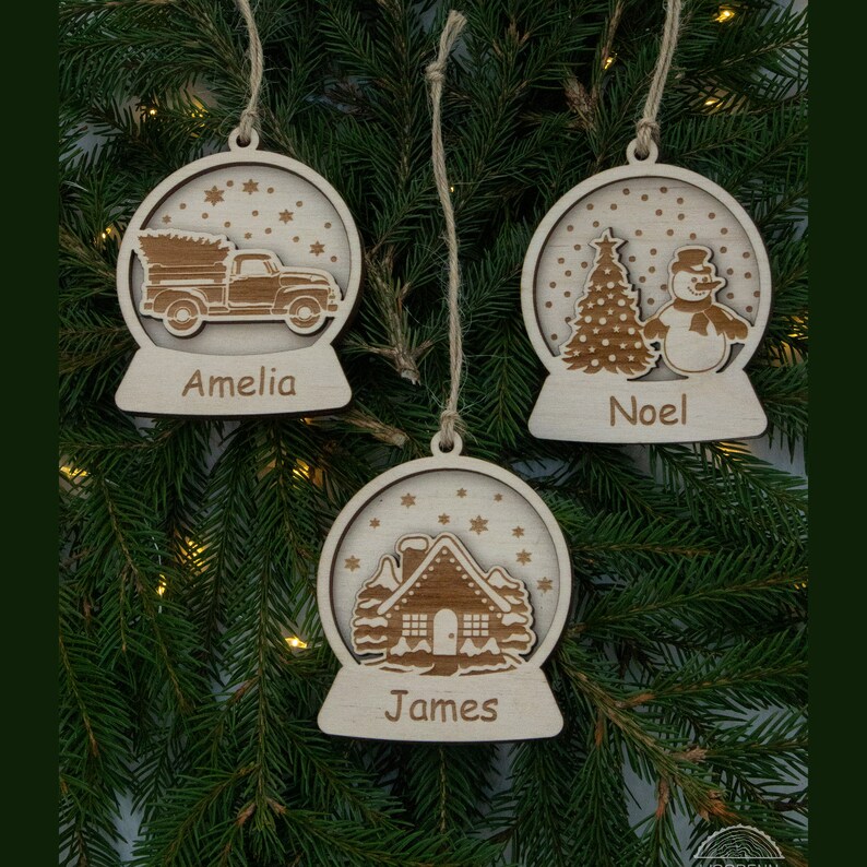 Pu&ograve; includere: Tre addobbi natalizi in legno a forma di palla di neve con diverse scene al loro interno. Gli addobbi sono personalizzati con i nomi Amelia, Noel e James.