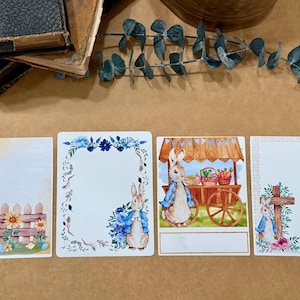 May include: Four printable cards featuring a watercolor illustration of a bunny rabbit. The cards have a white background with a blue floral border, a brown wooden cart with a bunny rabbit, a white background with a floral border, and a white background with a cross and floral border.