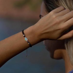 Puede incluir: Una pulsera de cordón negro con una pequeña piedra redonda azul claro y detalles plateados. La pulsera está en un brazo, con un fondo borroso de agua y tierra.