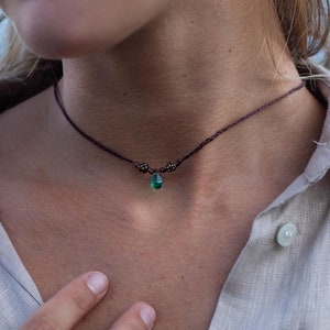 Op de afbeelding: Een donkerbruine koordketting met een kleine, druppelvormige, smaragdgroene hanger. De ketting heeft twee kleine decoratieve kralen bij de hanger. De persoon draagt een beige overhemd met knopen en een gouden ring.