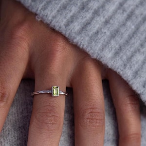 May include: Two silver rings with green gemstones. The ring on the index finger has a single, rectangular, light green stone. The ring on the pinky finger has a row of smaller, round, dark green stones.