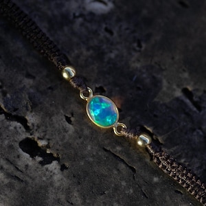 May include: A bracelet featuring a central oval-shaped opal gemstone with iridescent blue and green flashes, set in a gold-toned metal bezel. The stone is flanked by two small gold-toned beads on a dark brown woven cord with a textured finish.